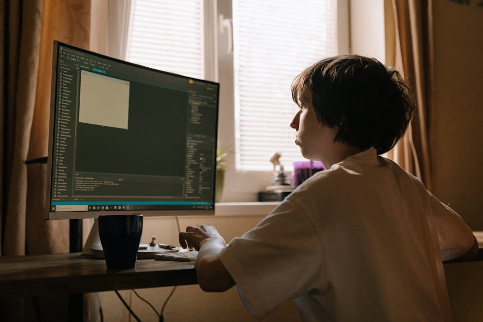 A person with short dark hair, wearing a light-colored shirt, is seated at a wooden desk indoors, working on a computer with a large monitor displaying code or a programming interface. The room has be