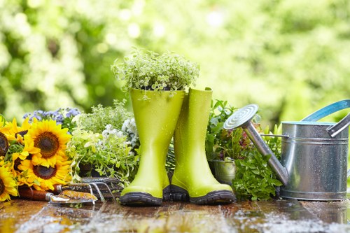 A garden scene featuring a weathered wooden table with gardening tools and decorative elements. On the left side, there are vibrant yellow sunflower flowers and smaller purple flowers arranged informally, adding colour contrast. In the centre, a pair of bright green rubber boots serve as planters, filled with delicate white and green plants, creating a charming focal point. To the right of the boots, a familiar metal watering can with a long spout and a blue garden fork resting on the table are visible, indicating active outdoor garden care. The background reveals a lush, green outdoor environment with leafy trees and bushes softly blurred, suggesting a well-maintained garden space typical of Highbury. The surface of the table appears slightly damp, hinting at recent watering or rain, enriching the natural ambience. This image illustrates a typical outdoor gardening setup, emphasizing practical tools and decorative plant elements, relevant to professional garden maintenance services offered by Garden Maintenance Highbury in London.